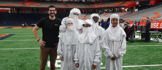 This is a photo of a group of students wearing clean suits, standing with a Micron employee and smiling at the camera.