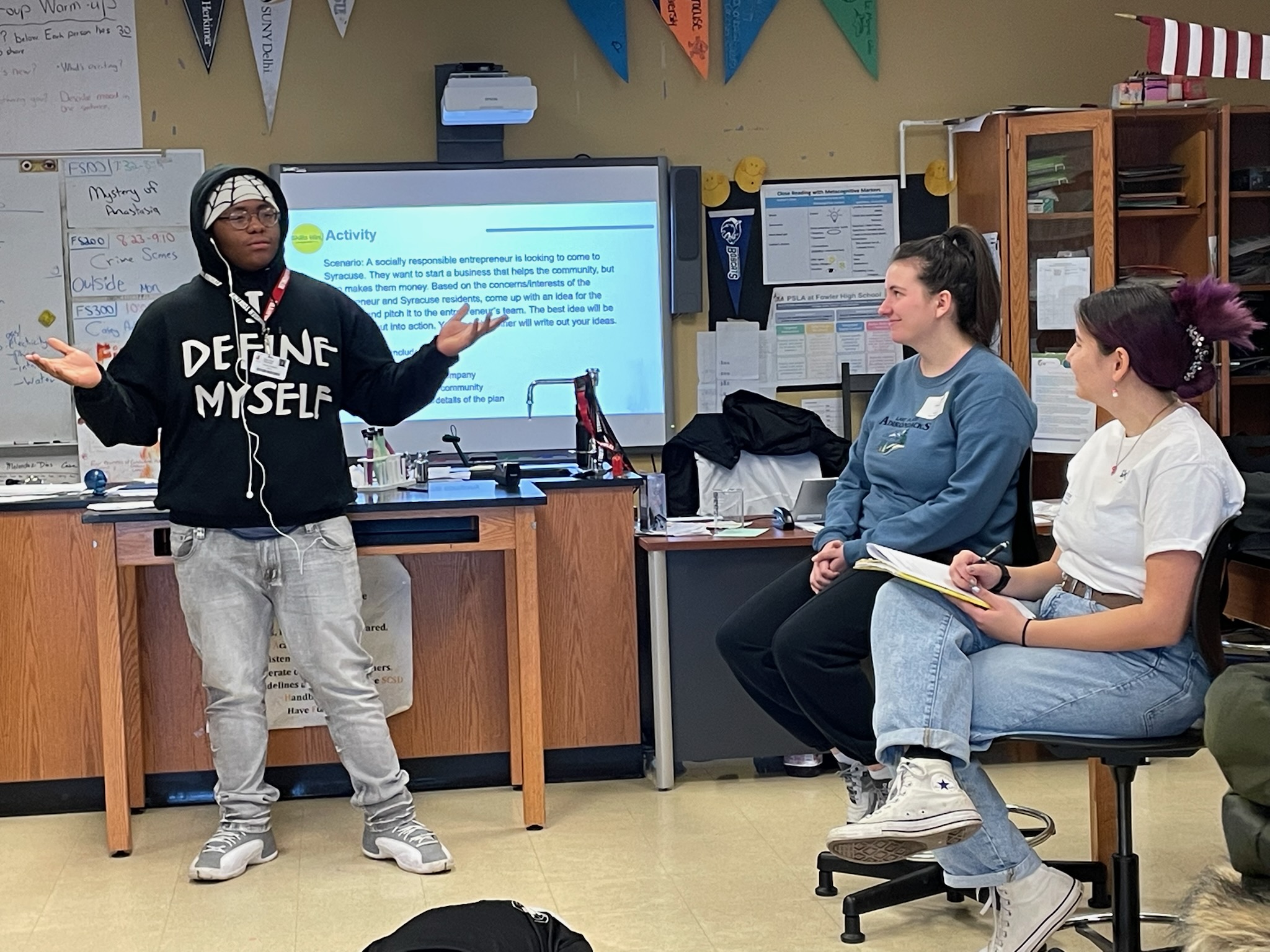 This is a photo of a PSLA at Fowler student presenting to his class, with two Syracuse University student mentors looking on.