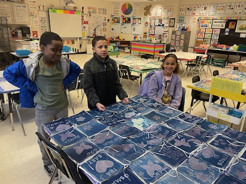 This is a photo of three students standing around a quilt on a table in their art classroom.