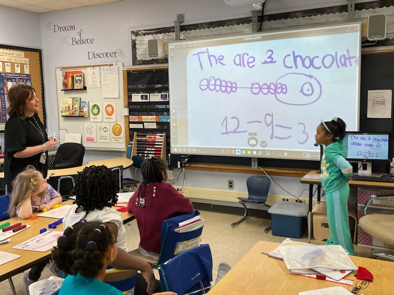 This is a photo of a first grade girl standing in front of the classroom screen projector showing her work to her classmates, who are looking on from their desks.
