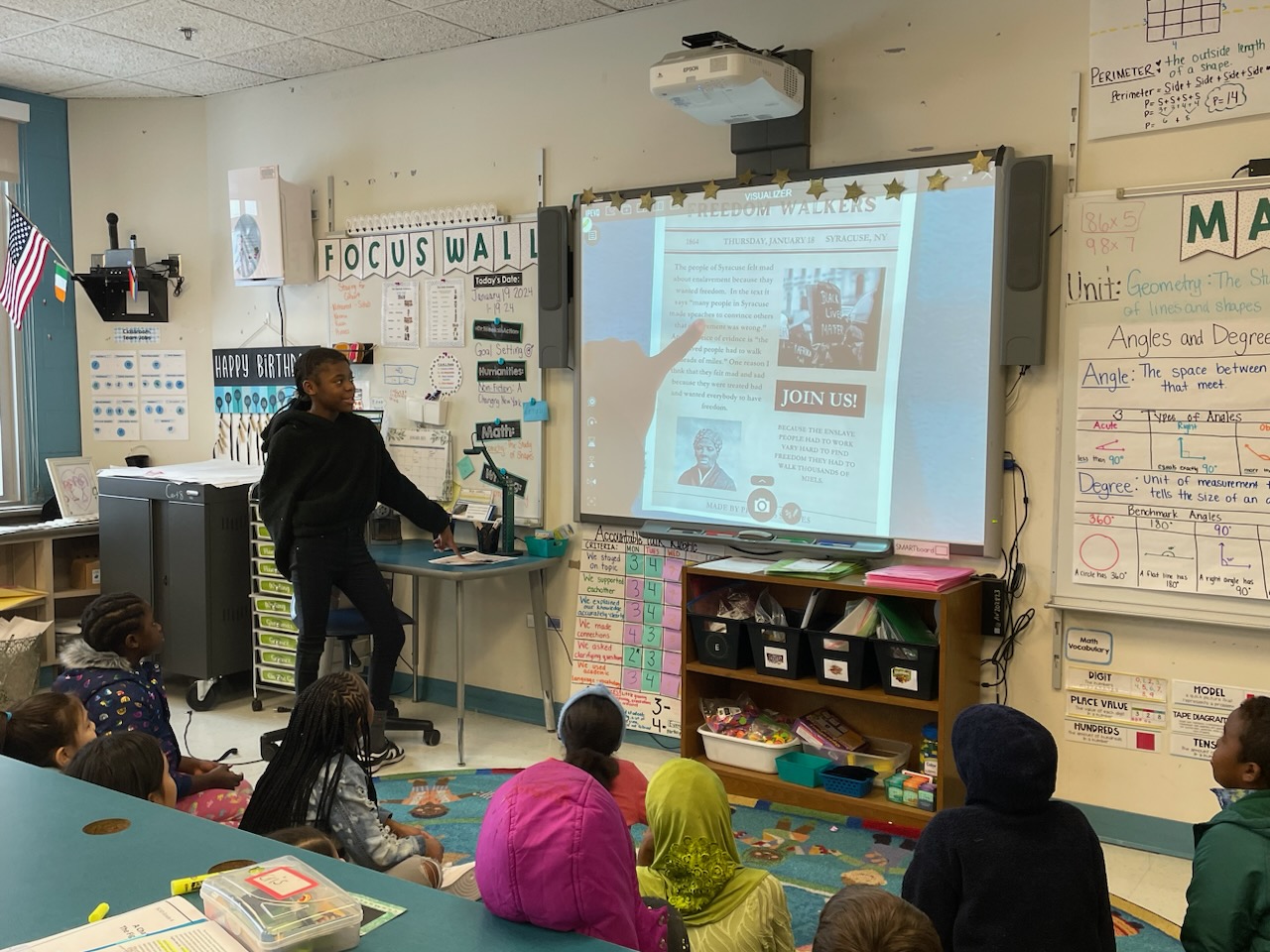 This is a photo of a Dr. Weeks 4th grade student standing in front of her class, pointing to her project displayed on the smart board in front of her.