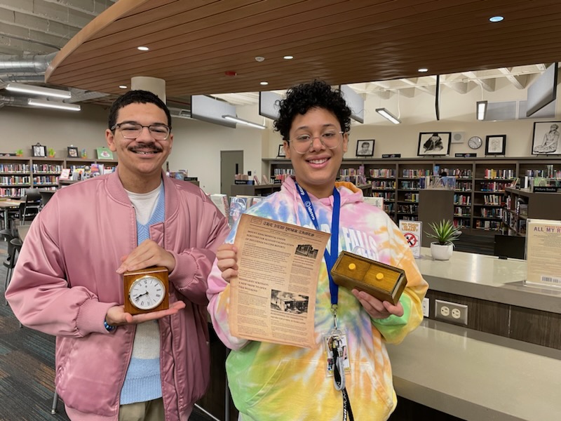 This is a photo of two students standing in the Henninger library, smiling at the camera, each holding an artifact they discovered during a library scavenger hunt.