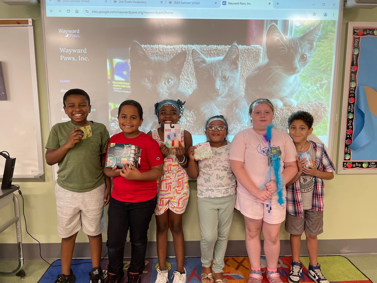 This is a photo of six Bellevue summer school students standing in front of a photo of kittens, each holding a cat toy or supply.