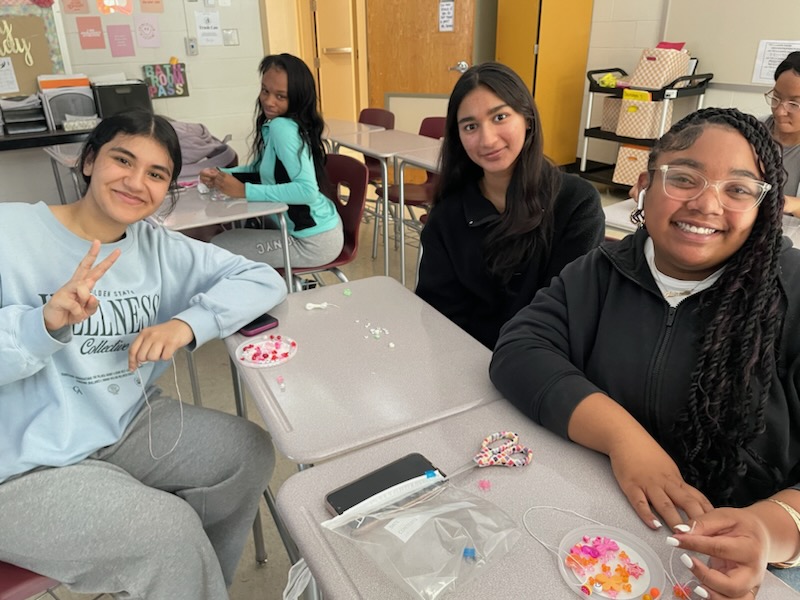 This is a photo of four students in Corcoran's fashion club sitting at desk making bead bracelets and smiling at the camera.