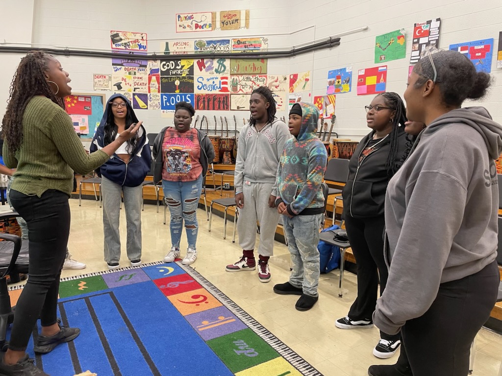 This is a photo of a group of Corcoran students, standing in a half circle in their chorus room and singing.