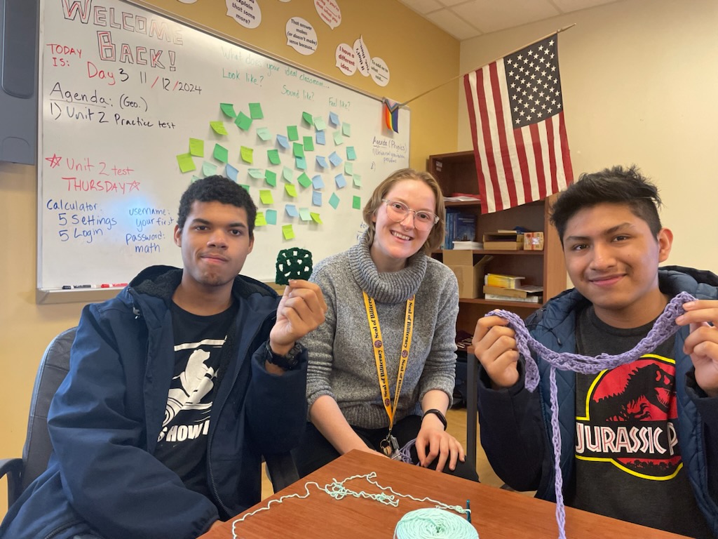 This is a photo of two students, holding knitting they have started, smiling with a teacher.