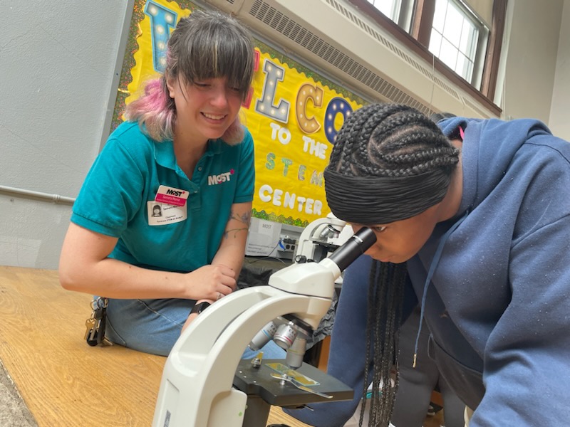 This is a photo of a Brighton Academy student peering into a microscope with a female volunteer looking on.