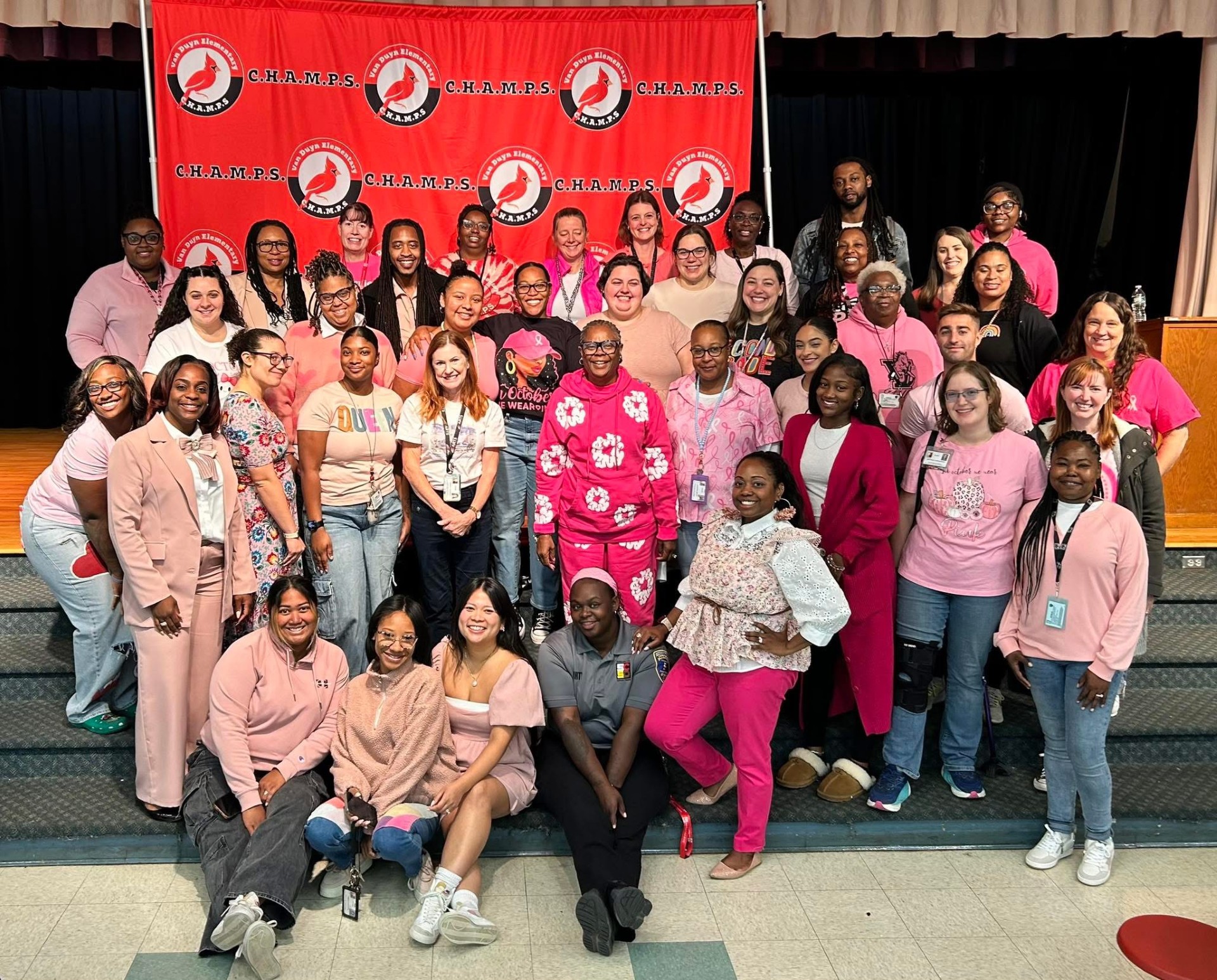 This is a photo of staff from Van Duyn Elementary School, wearing pink while standing on the steps of the school's stage.